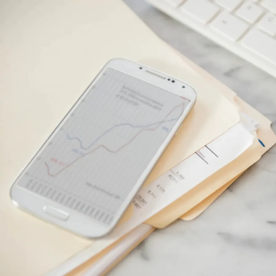 Ein Smartphone mit einem Liniendiagramm liegt auf ausgedruckten Finanzdokumenten auf einem Marmorschreibtisch, im Hintergrund sind eine weiße Tastatur und eine Brille zu sehen.