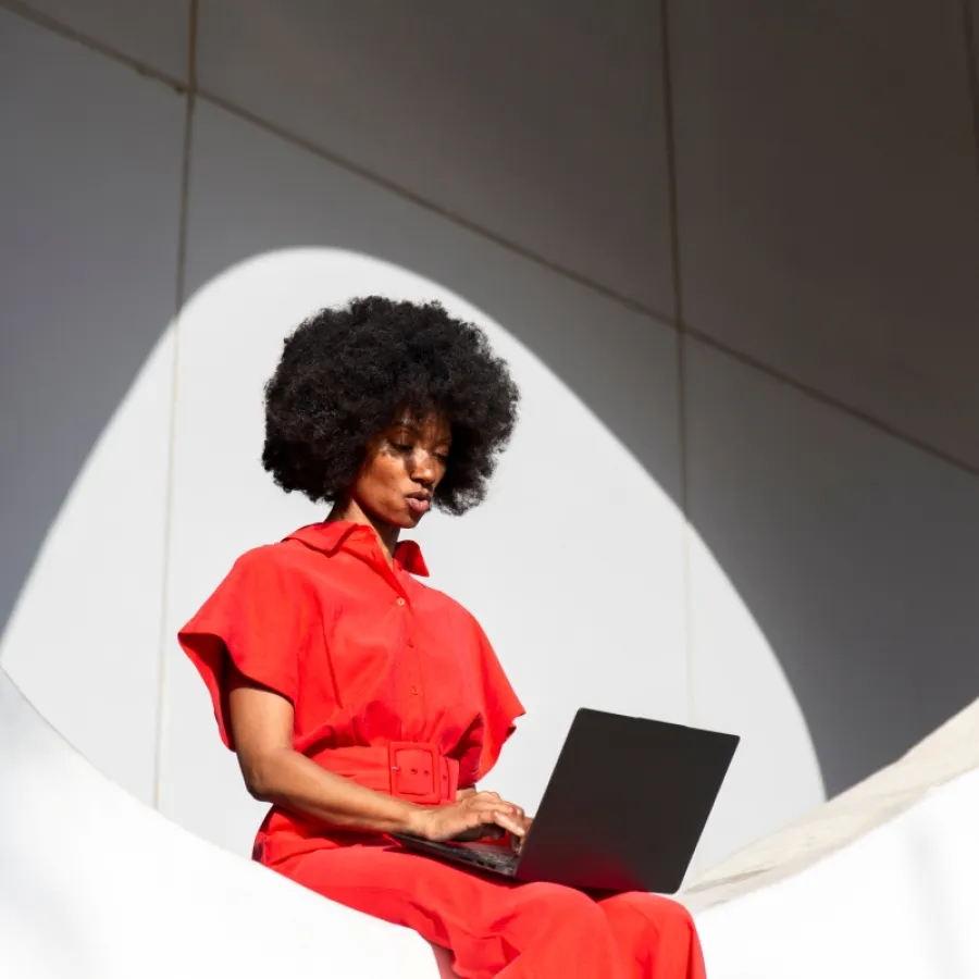 Eine Frau im roten Jumpsuit mit Afro-Frisur sitzt an einer weißen Wand und nutzt einen Laptop.