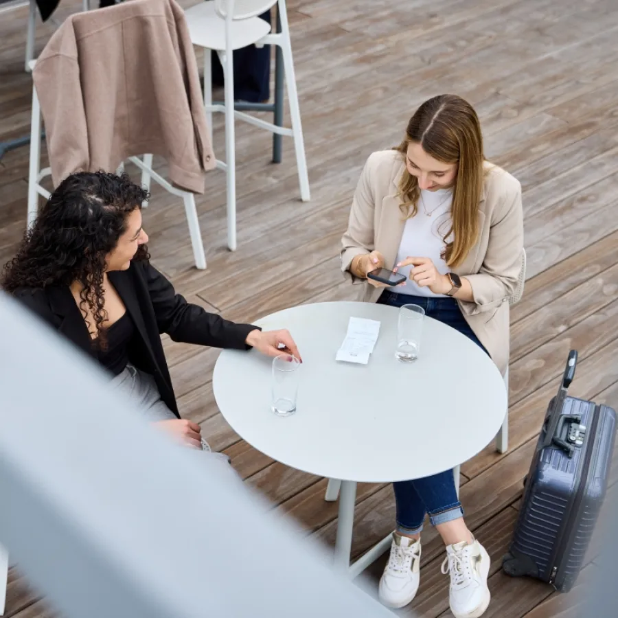 Zwei Business-Profis bei einem Gespräch im Außenbereich, möglicherweise in einem Café oder Besprechungsbereich, mit einem Koffer, der auf eine Geschäftsreise hinweist.